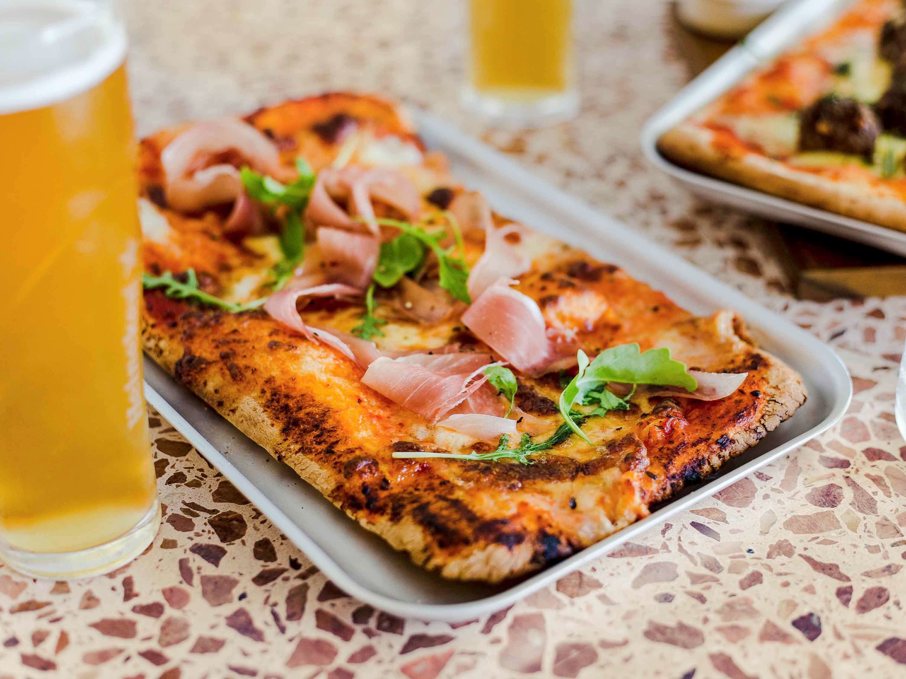 Pizza, jossa on päällä ilmakuivattua kinkkua ja vihreitä kasviksia, tarjoiltuna tarjottimella.
