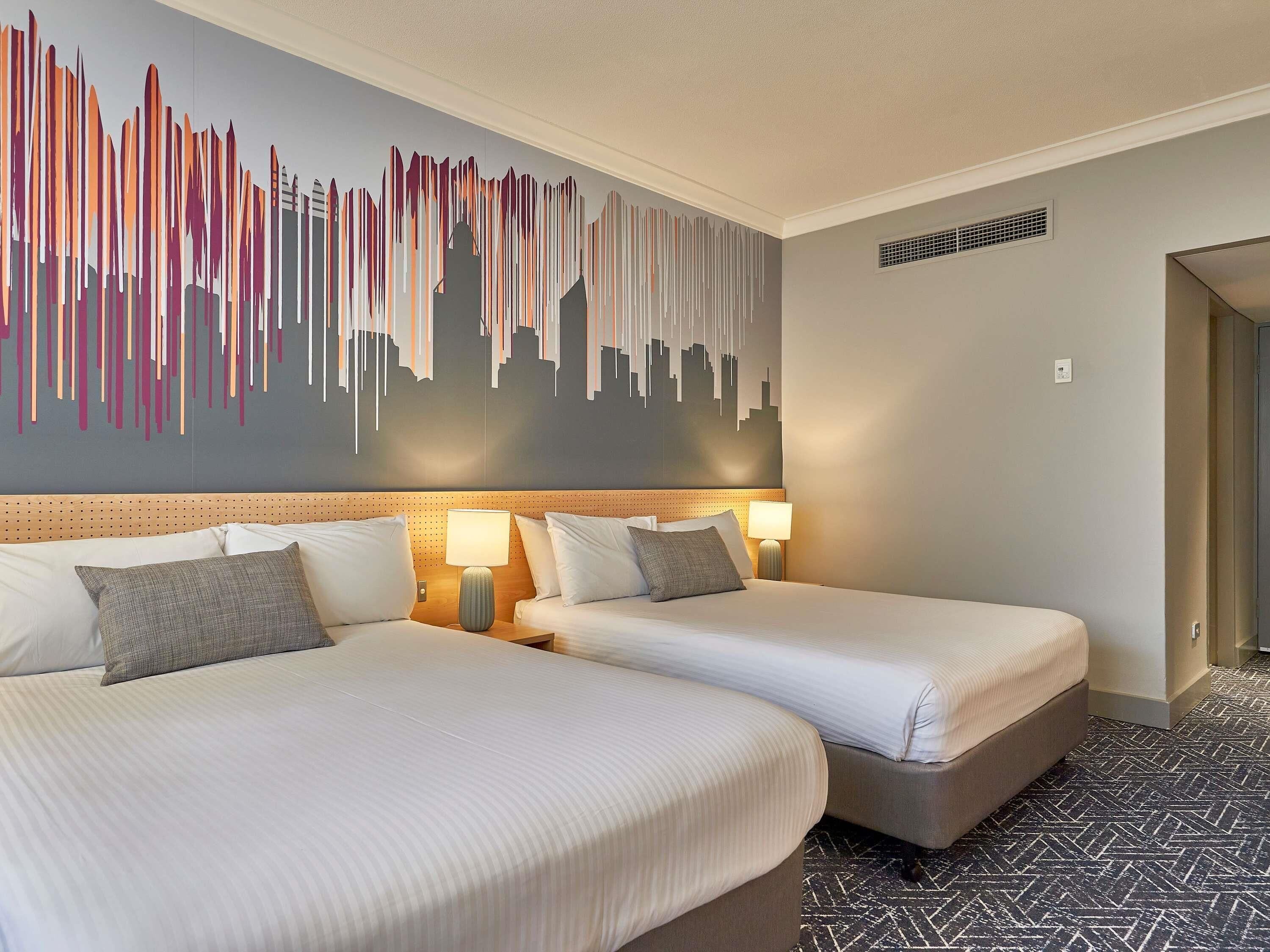 Habitación de hotel con dos camas, un cuadro de arte con vista urbana, cabecera de madera y suelo estampado.