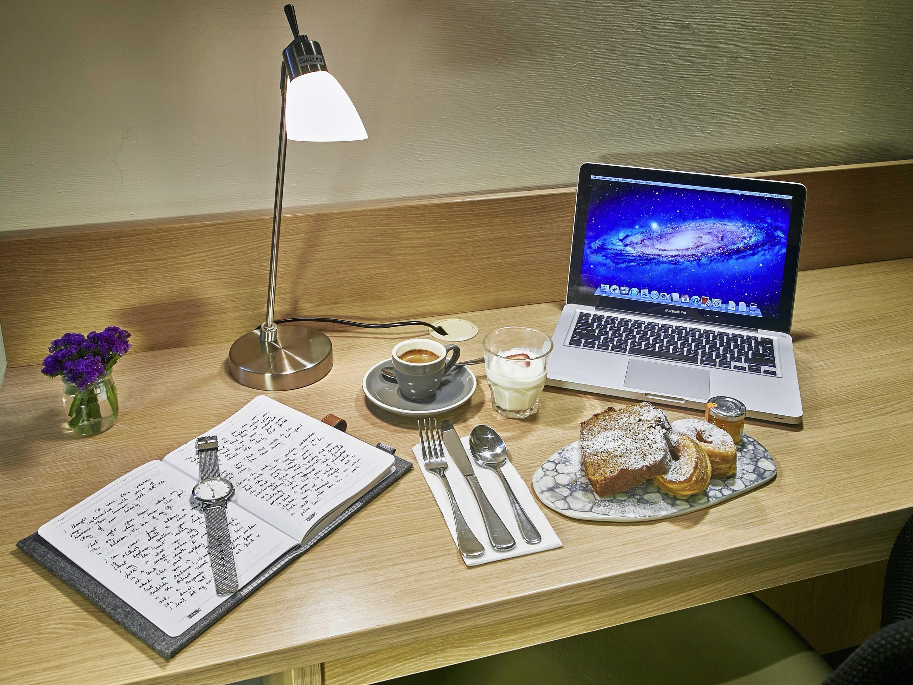A wooden desk with a laptop, a cup of coffee, a glass of milk, a plate of pastries, a notebook, a pen, a watch, and a vase with purple flowers.