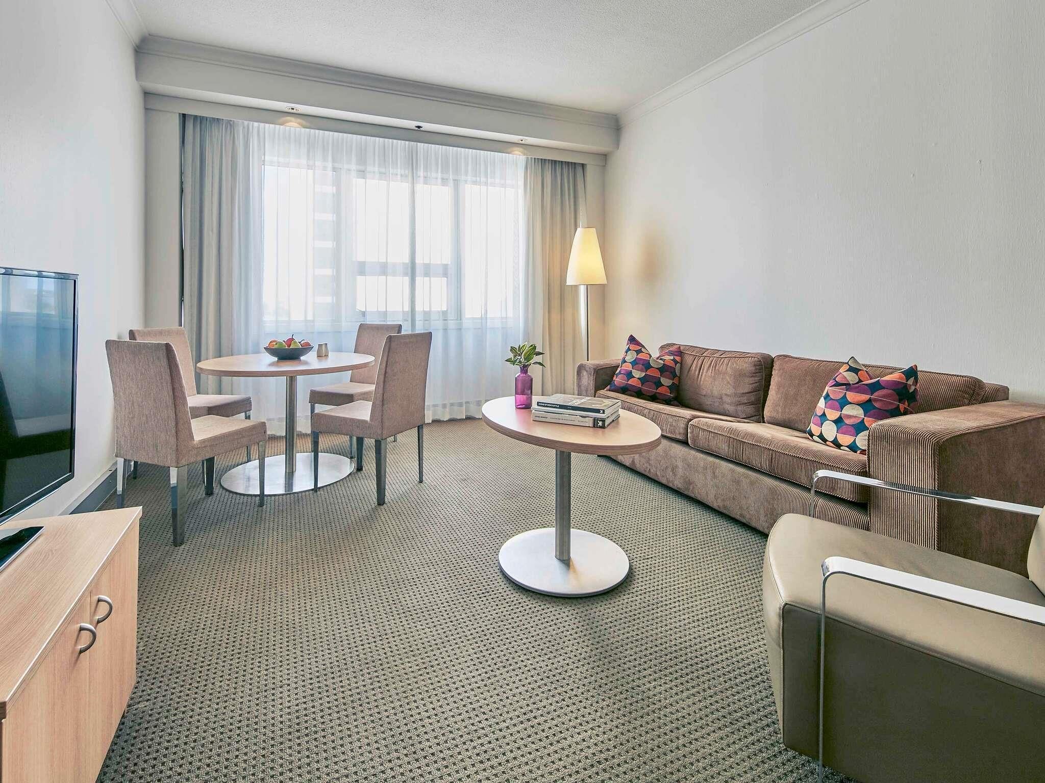 A hotel room with a sofa, a dining table with chairs, a TV, and a lamp.