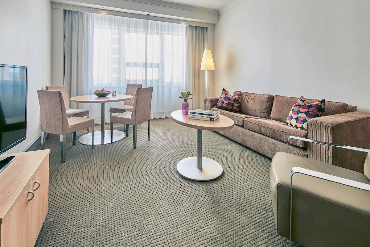 A hotel room with a sofa, a dining table with chairs, a TV, and a lamp.