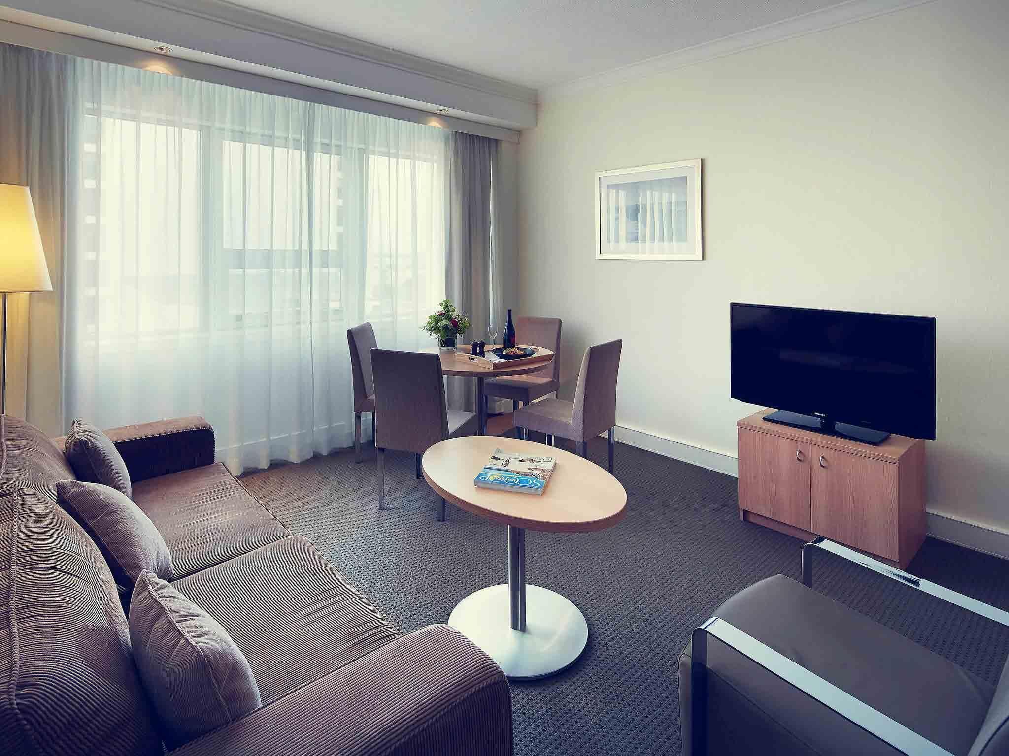 A hotel room with a sofa, a small round table, a television, and a dining area with chairs and a table.