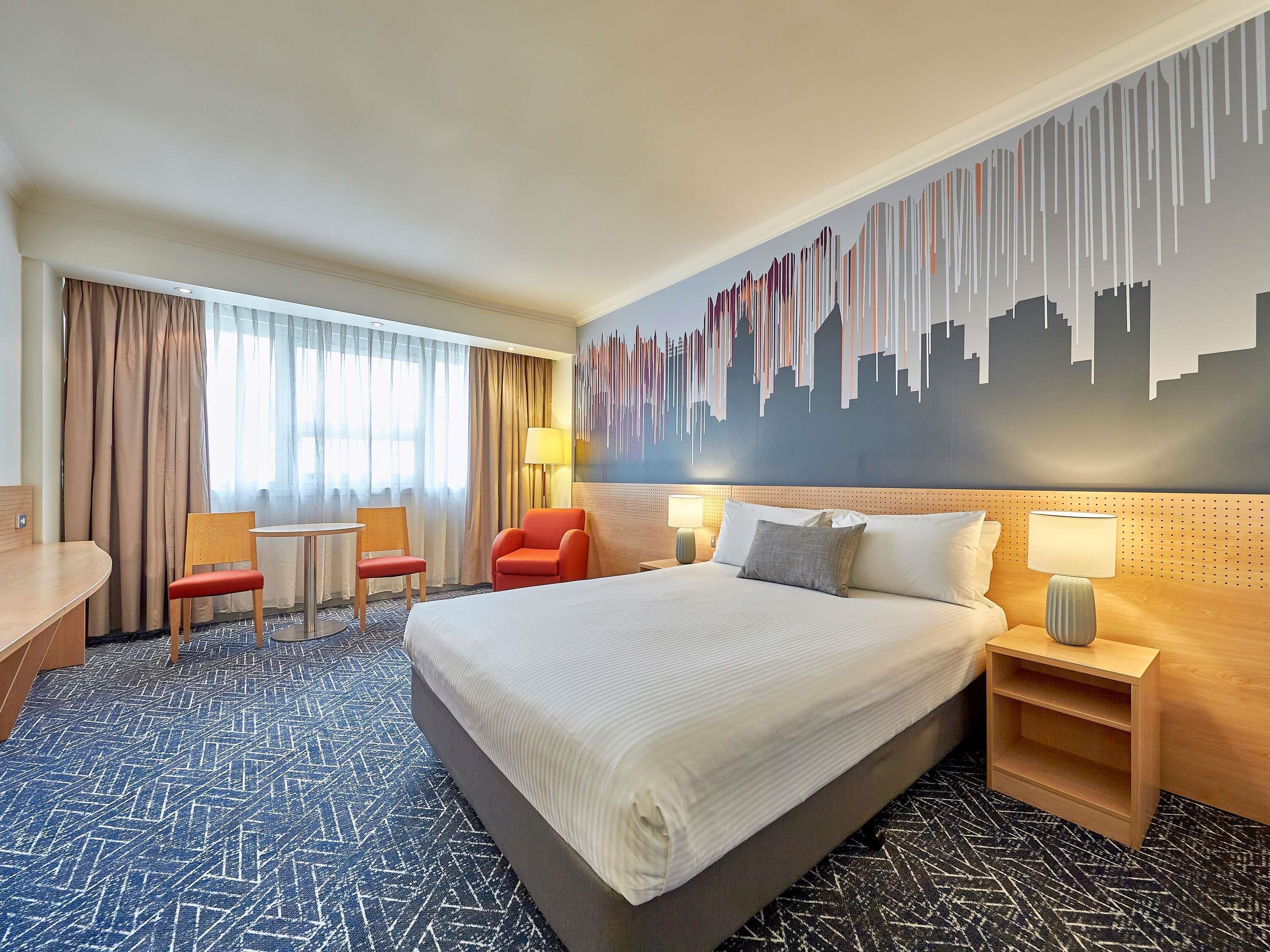 A hotel room with a large bed, two chairs, a desk, and a cityscape mural on the wall.