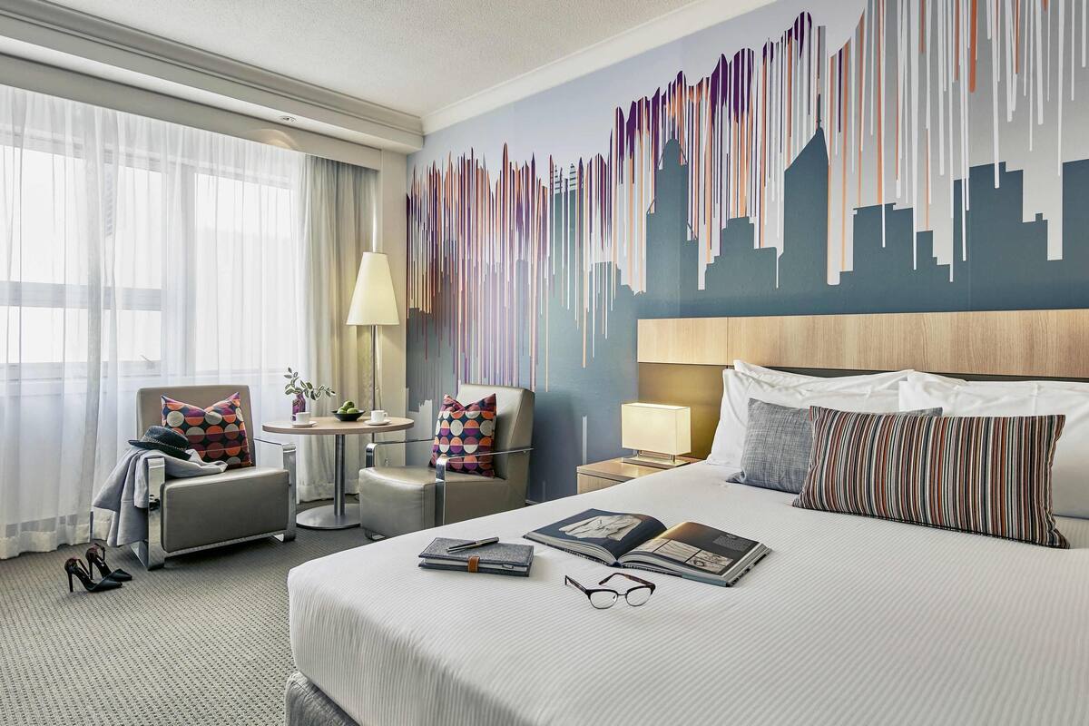 Ein Hotelzimmer mit einem großen Bett, zwei Sesseln, einem kleinen Tisch und einem Stadtbild-Wandgemälde.