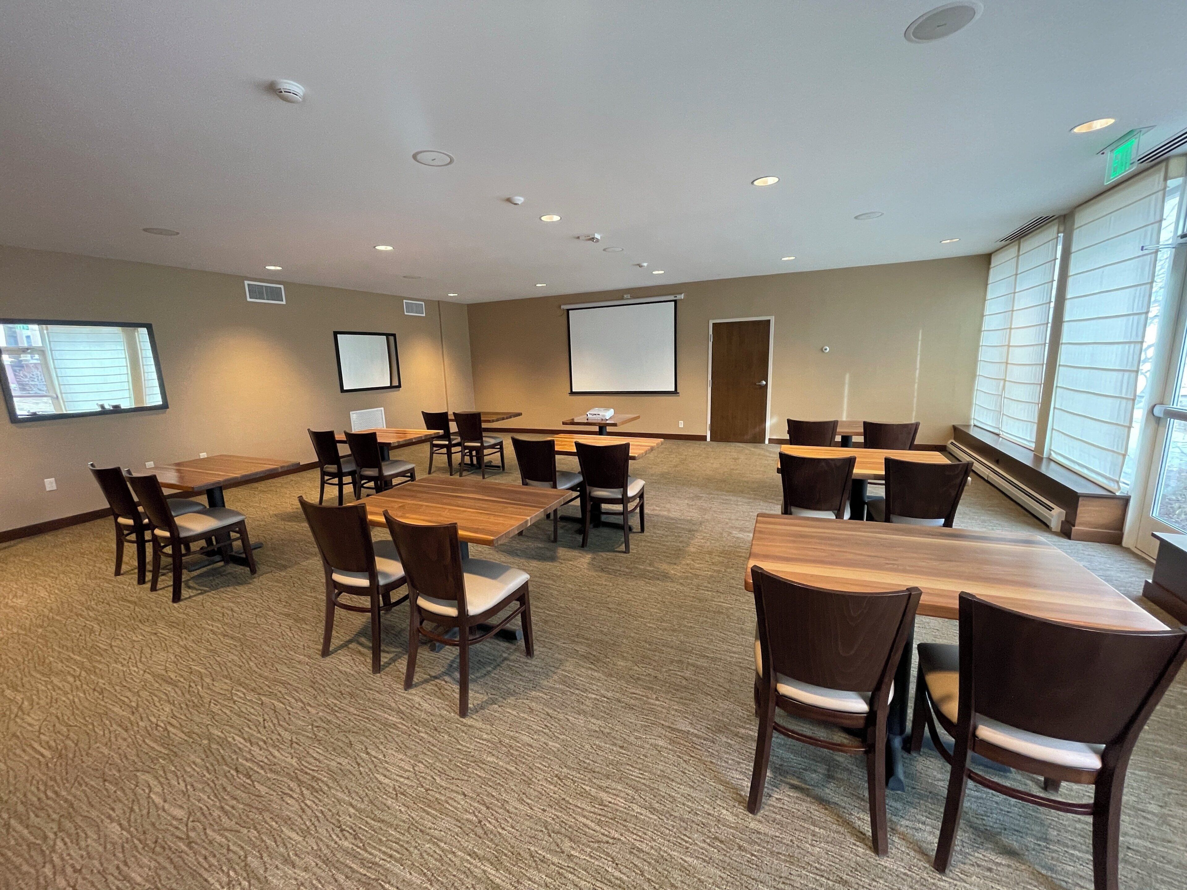 A conference room with multiple tables and chairs, a projector screen, and a large window with blinds.