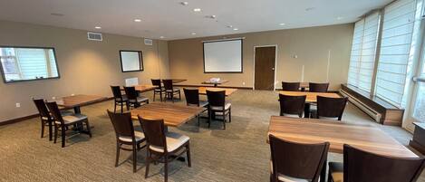 A conference room with multiple tables and chairs, a projector screen, and a large window with blinds.