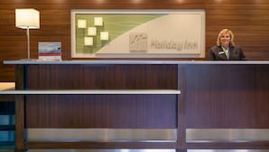 A reception desk with a wooden panel design, a framed sign with the name 'Holiday Inn', and a lamp to the left. - Holiday Inn Bangor by IHG (Bangor)