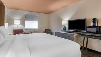 Habitación de hotel con una cama grande, un escritorio con televisor y cafetera, una silla y una ventana con persianas.
