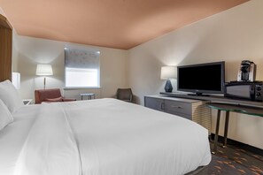 A hotel room with a large bed, a desk with a TV and coffee machine, a chair, and a window with blinds.