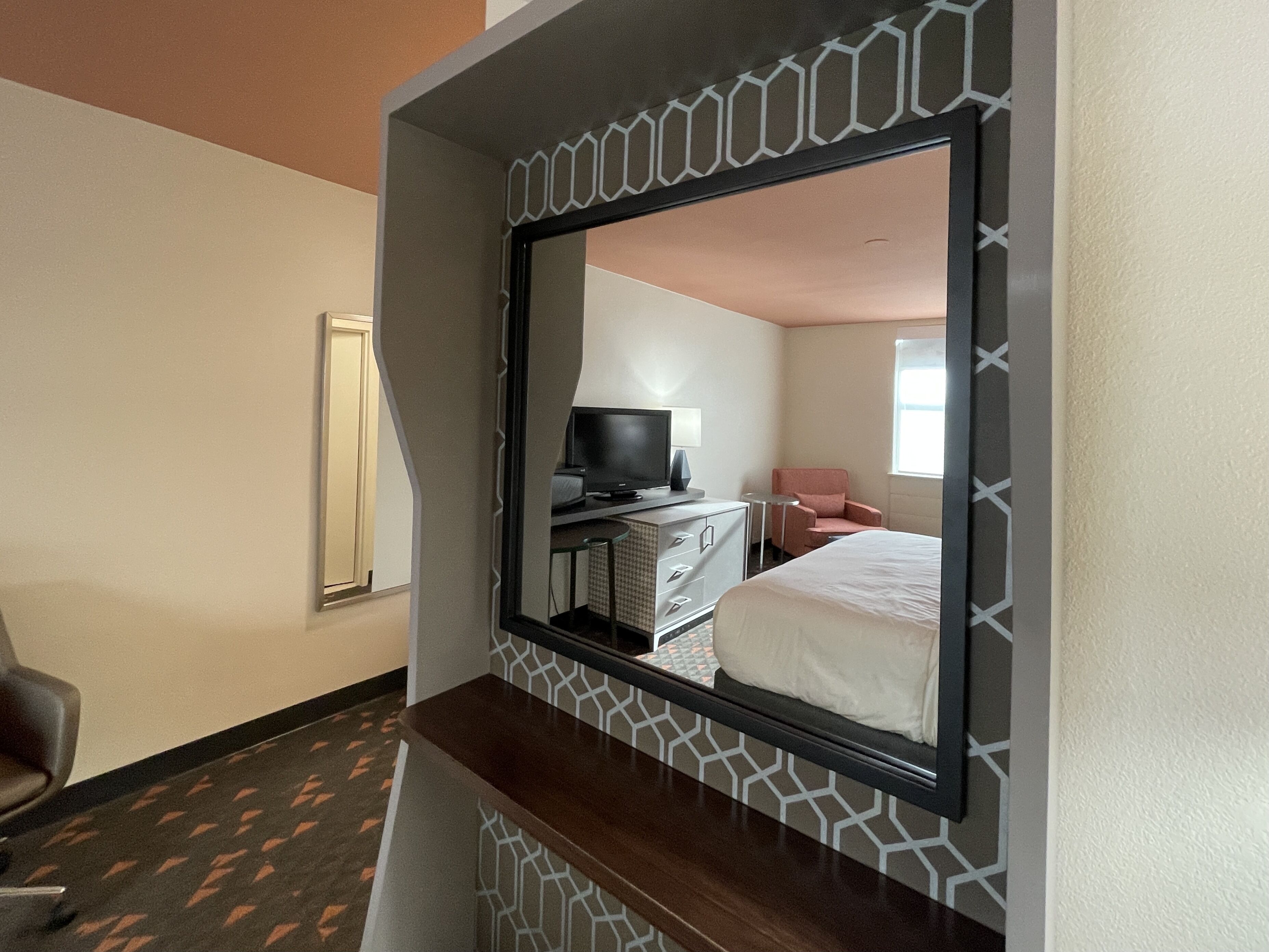 A hotel room with a large mirror, a bed, a desk with a computer, a chair, and a TV.
