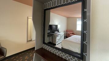 A hotel room with a large mirror, a bed, a desk with a computer, a chair, and a TV.