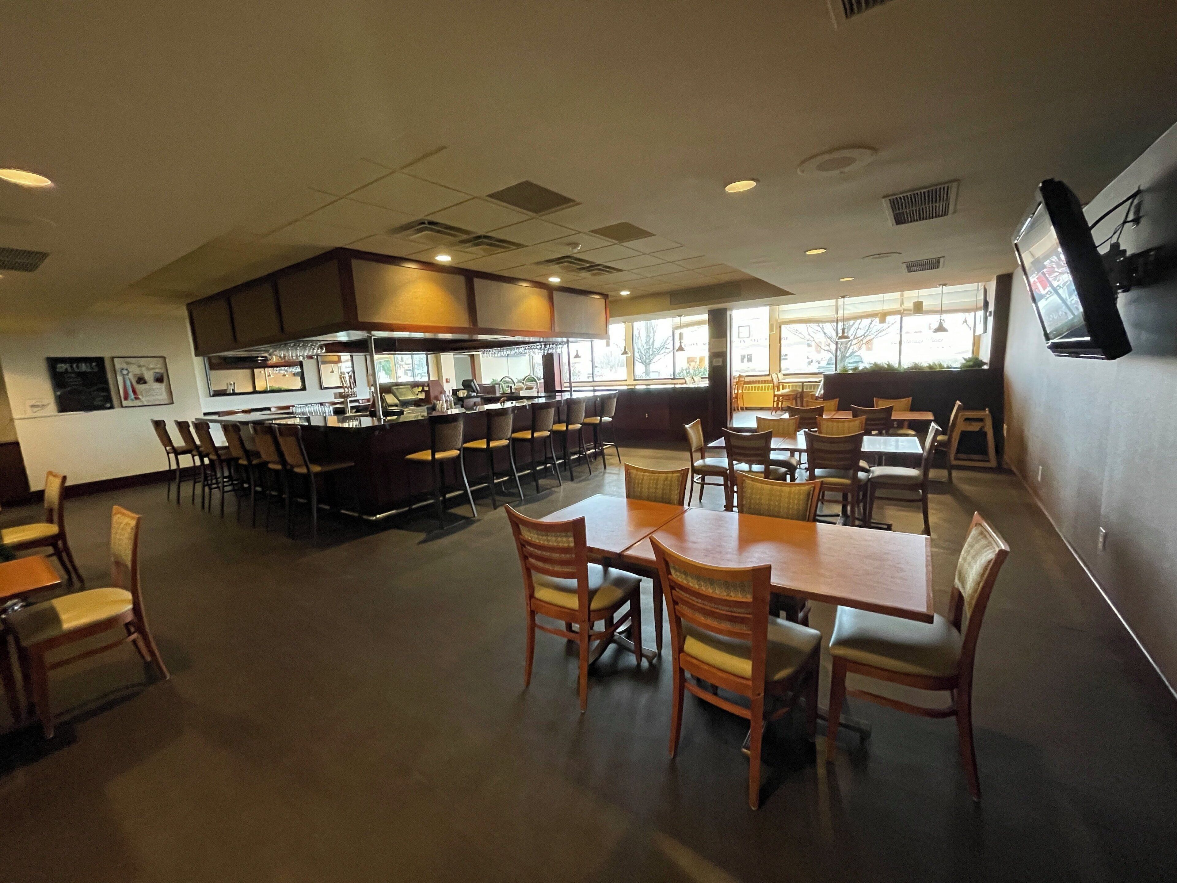 Um restaurante com área de bar, várias mesas e cadeiras, e cozinha ao fundo.