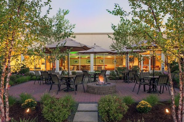 An outdoor patio area with tables and chairs, a fire pit, and umbrellas providing shade.