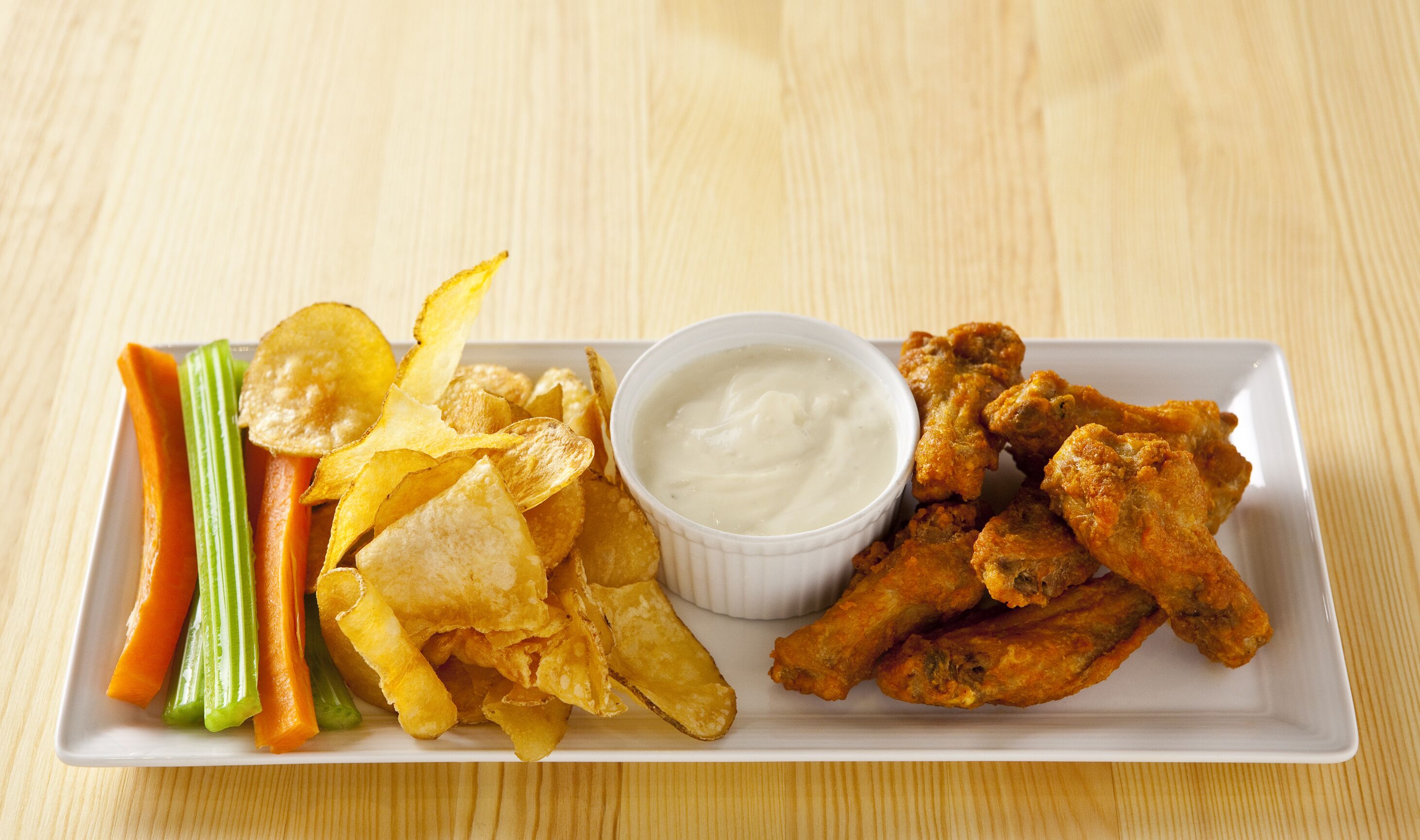 A rectangular white plate with a variety of food items including fried chicken wings, potato chips, celery sticks, carrot sticks, and a small bowl of dipping sauce.