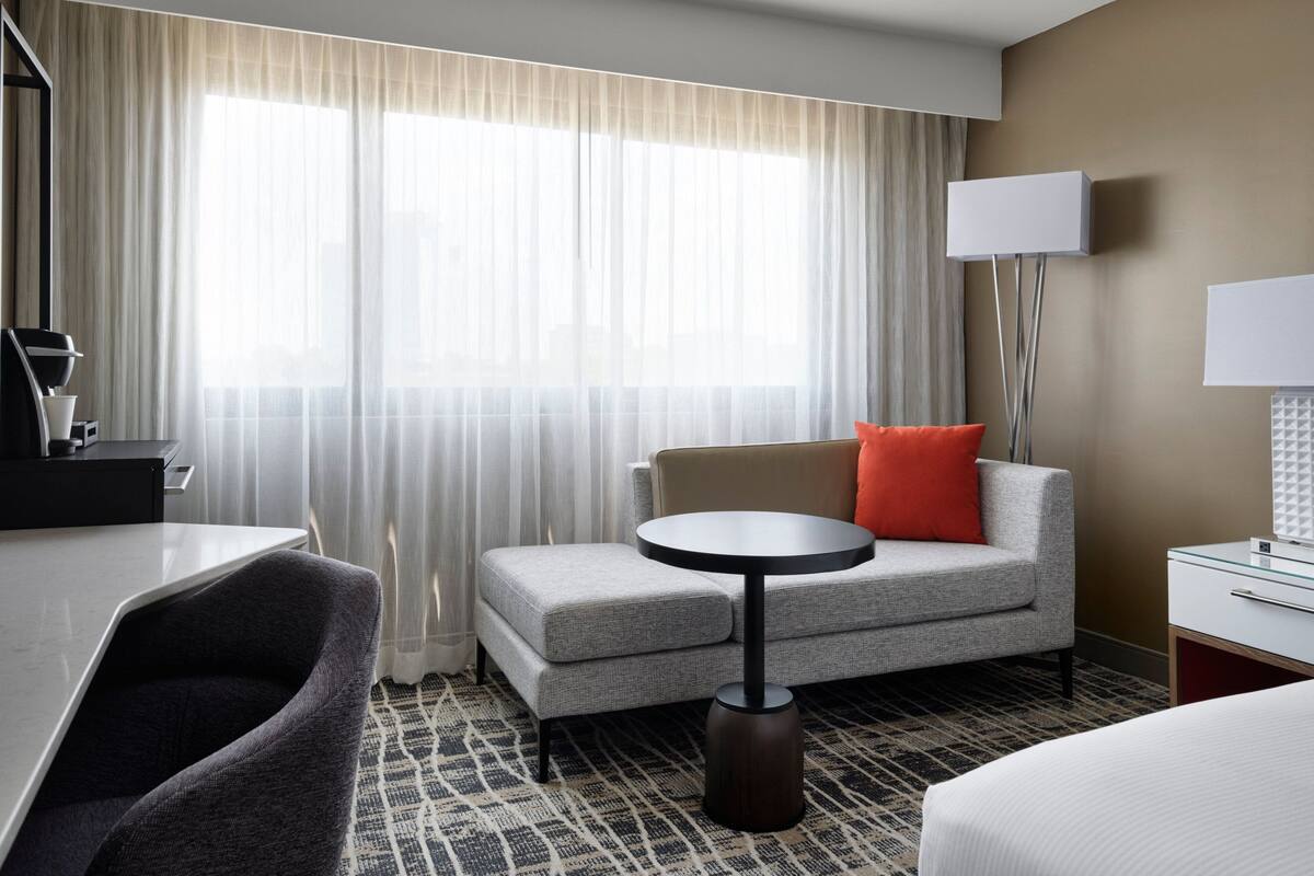 Una habitación de hotel con sofá, una mesita redonda pequeña, un escritorio con silla, cama y una ventana grande con cortinas translúcidas.
