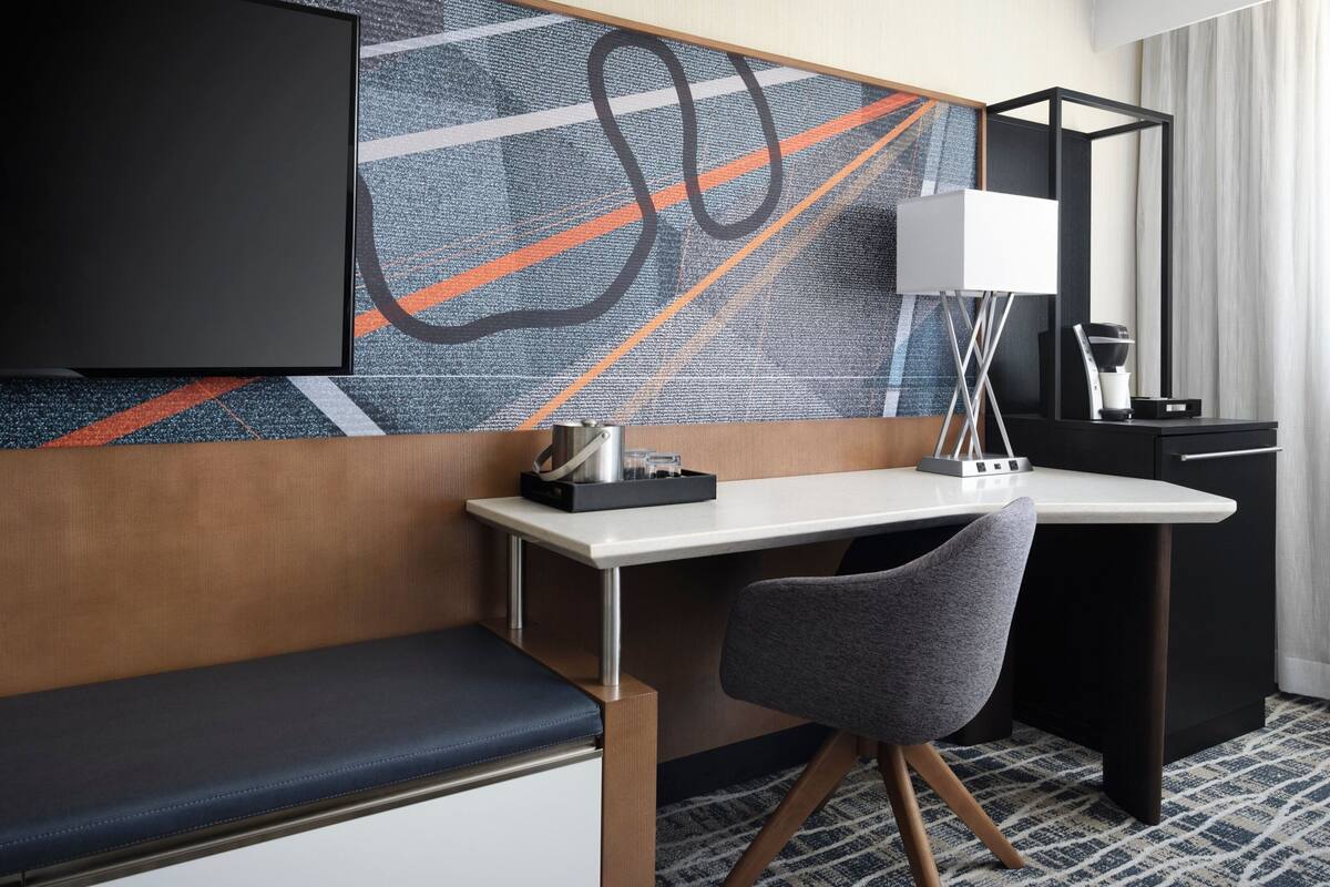 Habitación de hotel con escritorio, una silla, un banco, televisor, espejo y un cuadro abstracto en la pared.