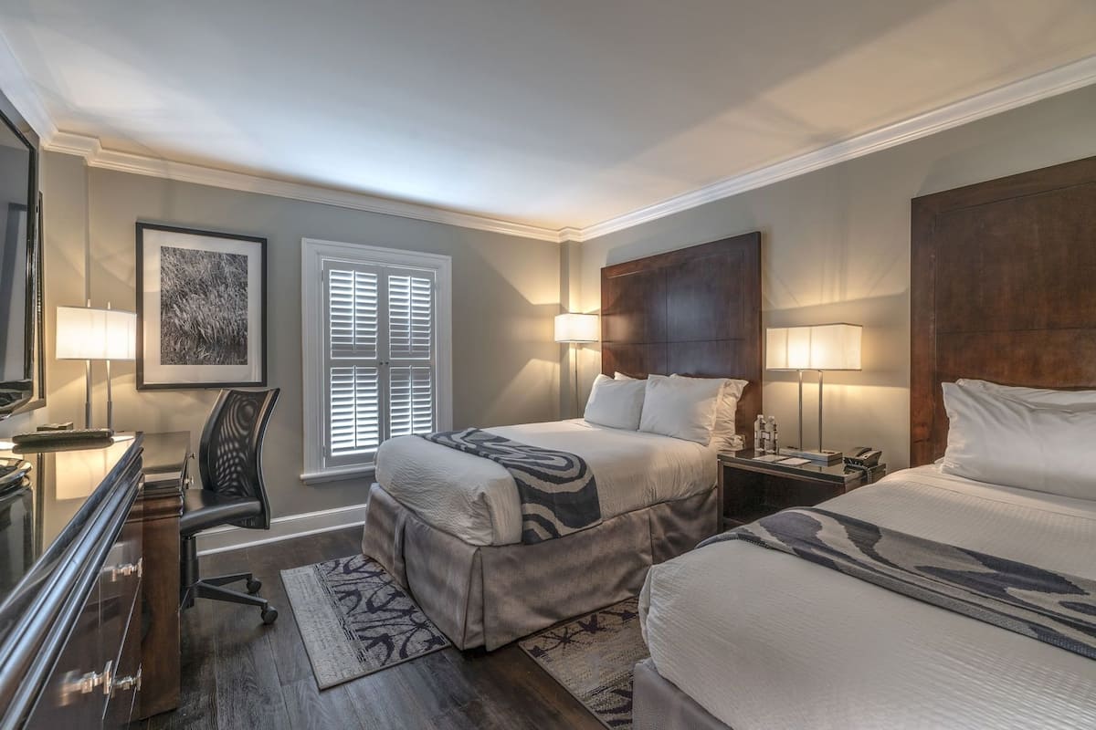 A hotel room with two beds, a desk, a chair, a desk lamp, a bedside table, a telephone, and a window with blinds.