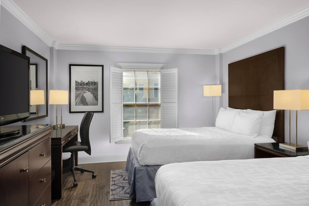 A hotel room with two beds, a desk, a chair, a television, and a window with shutters.