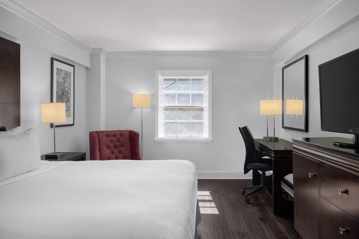 A hotel room with a bed, a desk, a chair, a dresser, a TV, and two lamps.