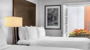 Camera d'albergo con un letto, un comodino, una lampada, una fotografia in bianco e nero incorniciata e una finestra che si affaccia su un muro di mattoni e fiori.