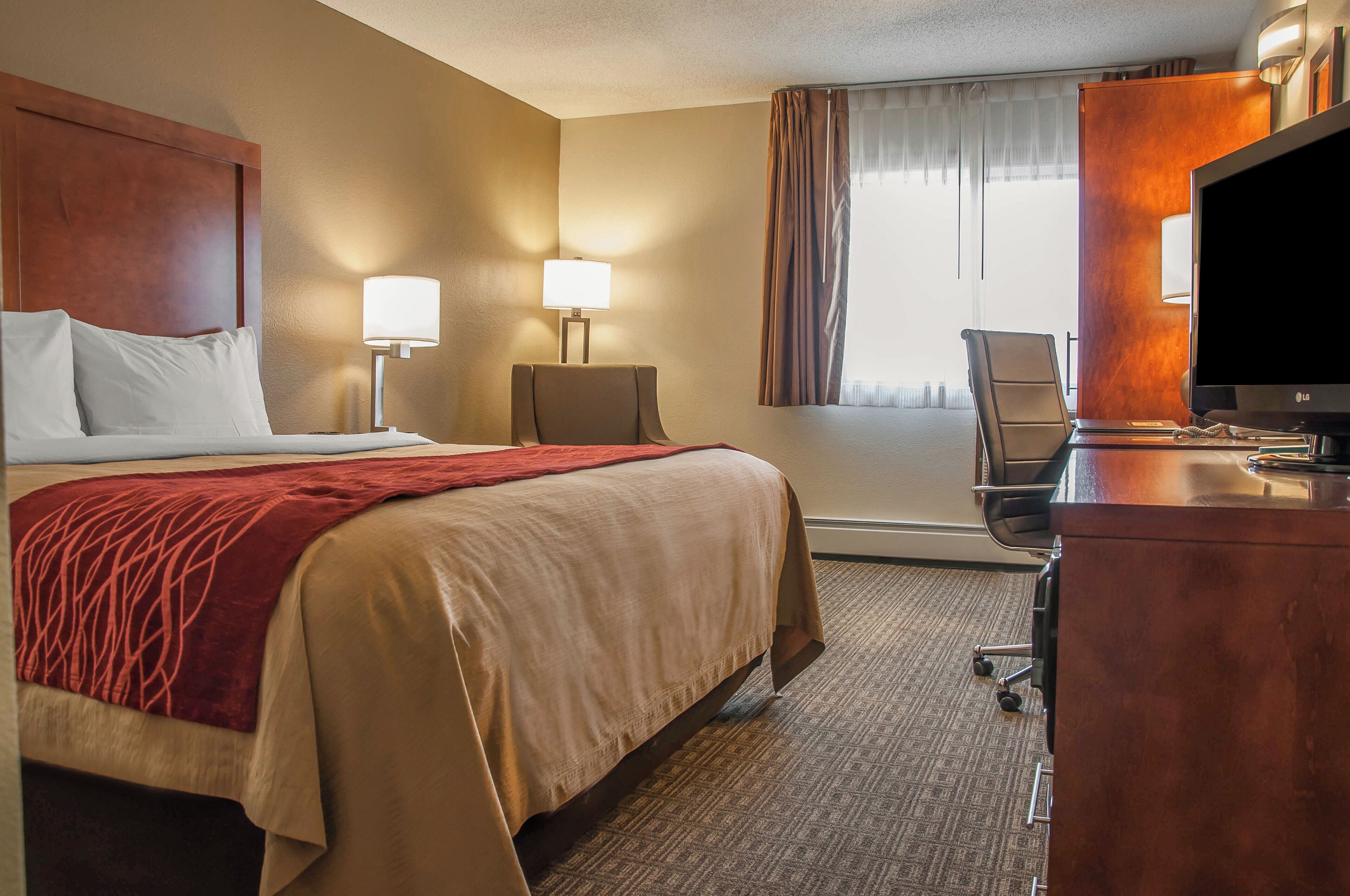 A hotel room with a large bed, two bedside lamps, a desk with a television, and a chair.
