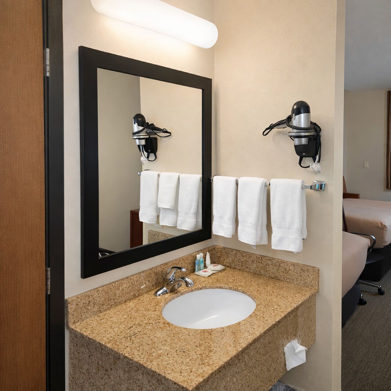 Casa de banho de hotel com lavatório de granito, um espelho grande e dois secadores de cabelo fixados na parede.