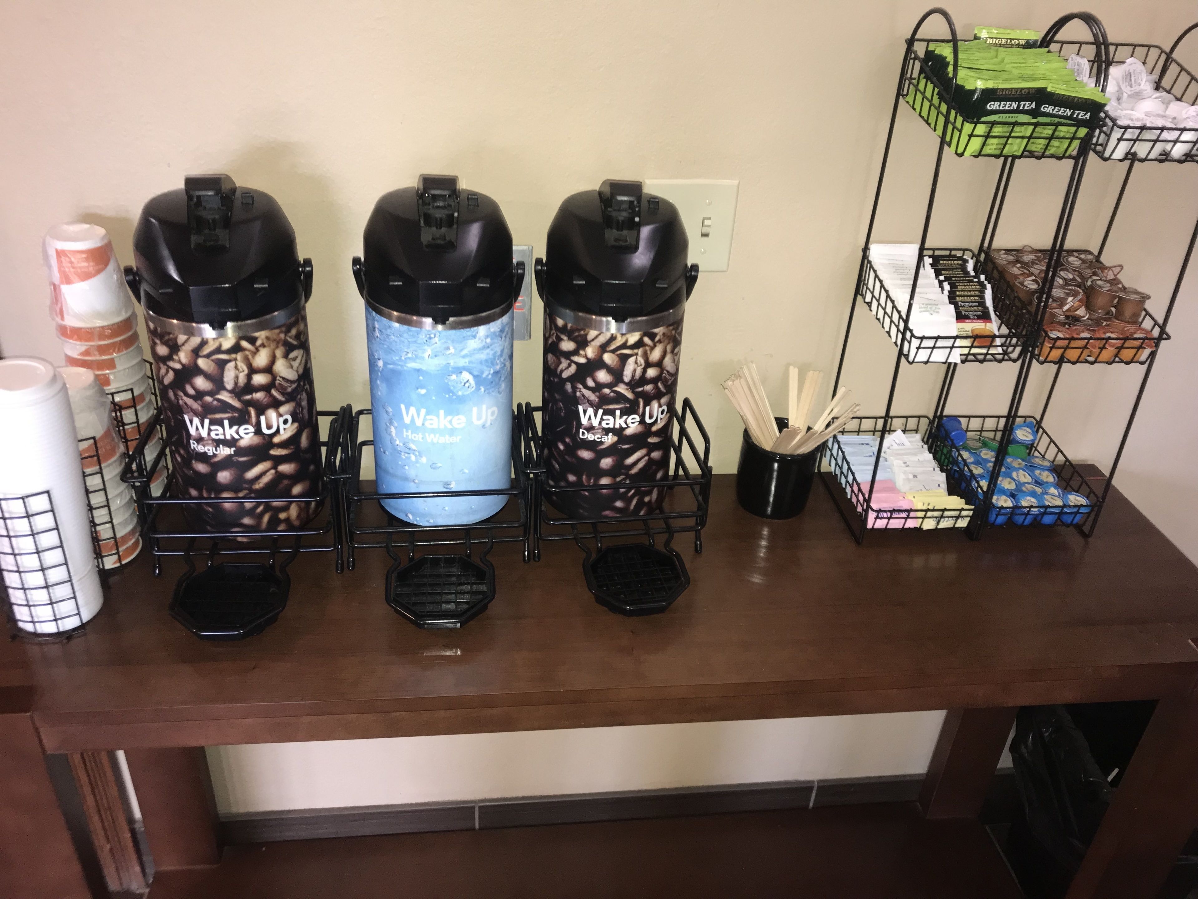 A coffee station with three coffee dispensers labeled "Wake Up" and a shelf with various coffee pods and utensils.