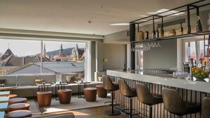 A modern bar area with a view of buildings through large windows, bar stools, and a variety of bottles and glasses on the shelves. - Garner Hotel Klagenfurt - Moser Verdino (Klagenfurt am Woerthersee)