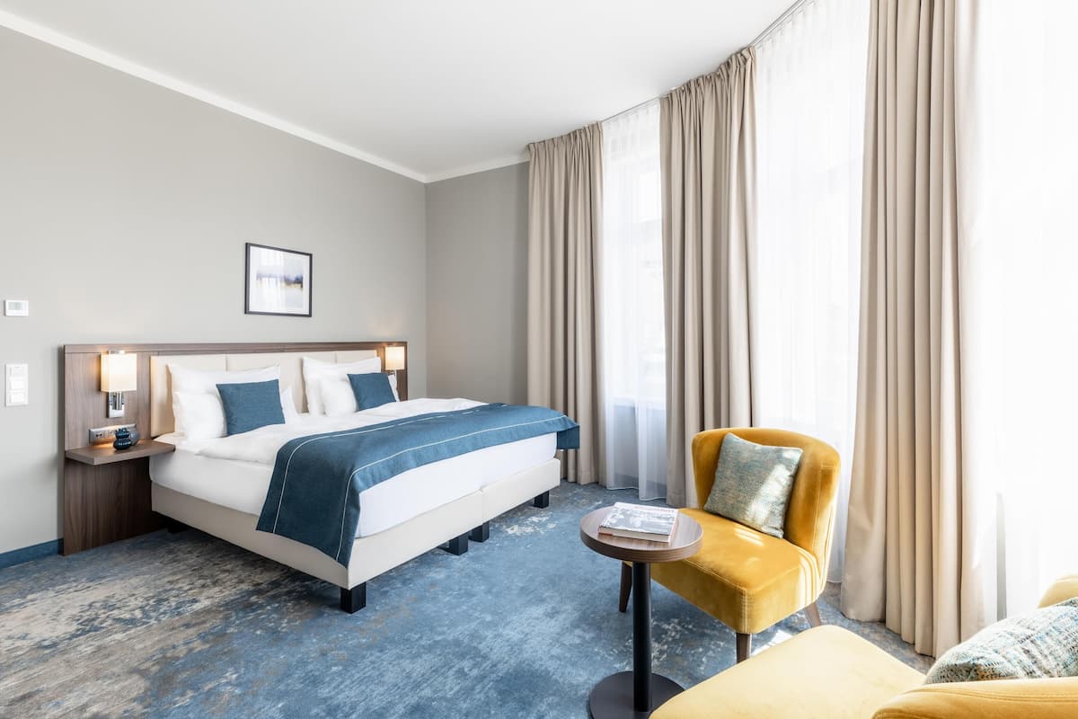 Una habitación de hotel con cama, una mesita redonda pequeña, un sillón amarillo y un ventanal con cortinas.