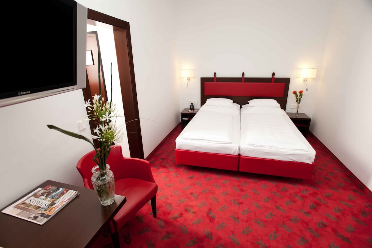 Habitación de hotel con alfombra roja, dos camas, un televisor, un escritorio con una revista, un florero con flores y una silla roja.