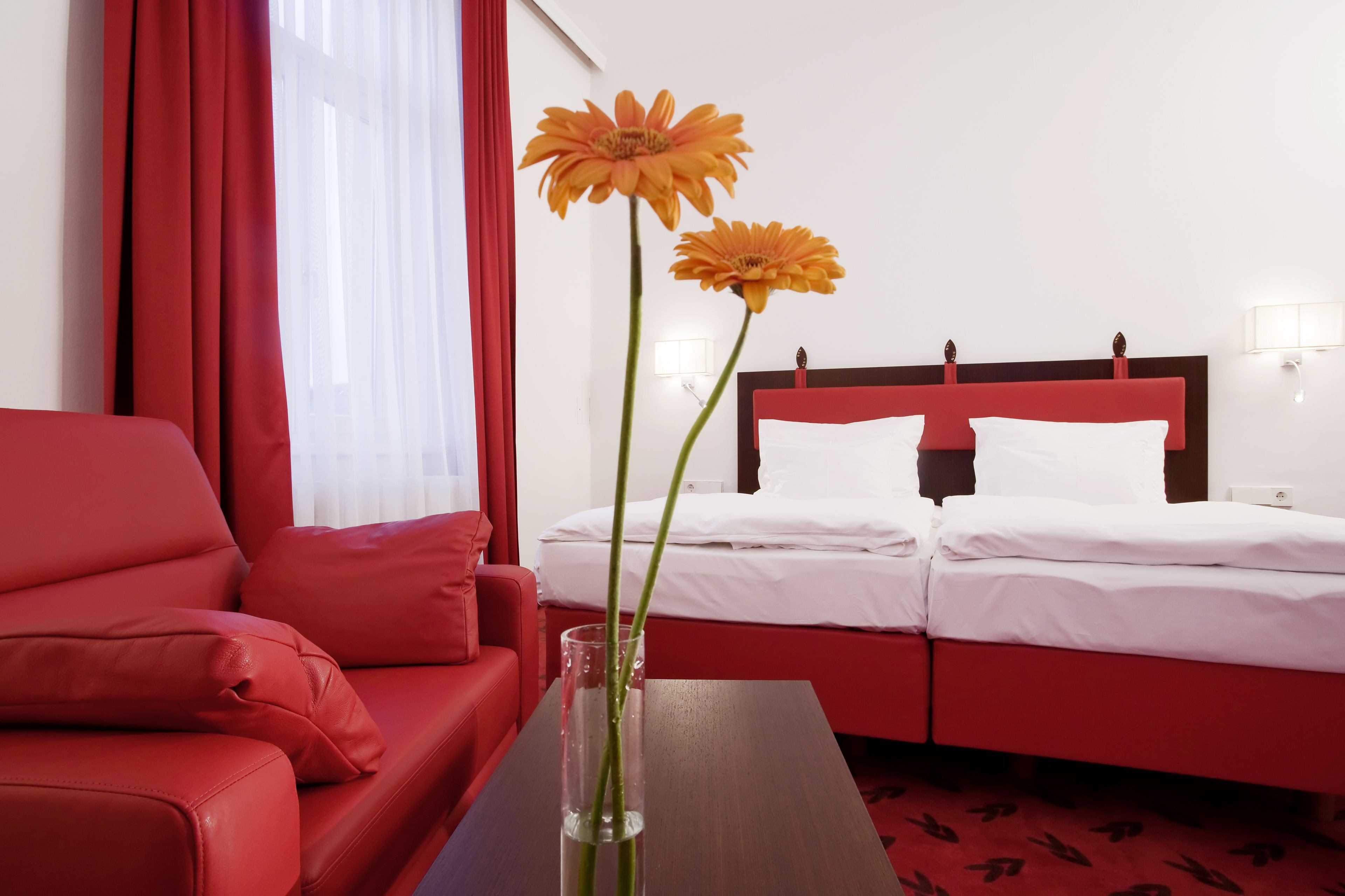 A hotel room with a red sofa, a bed with white bedding, and a vase with two orange flowers on a table.