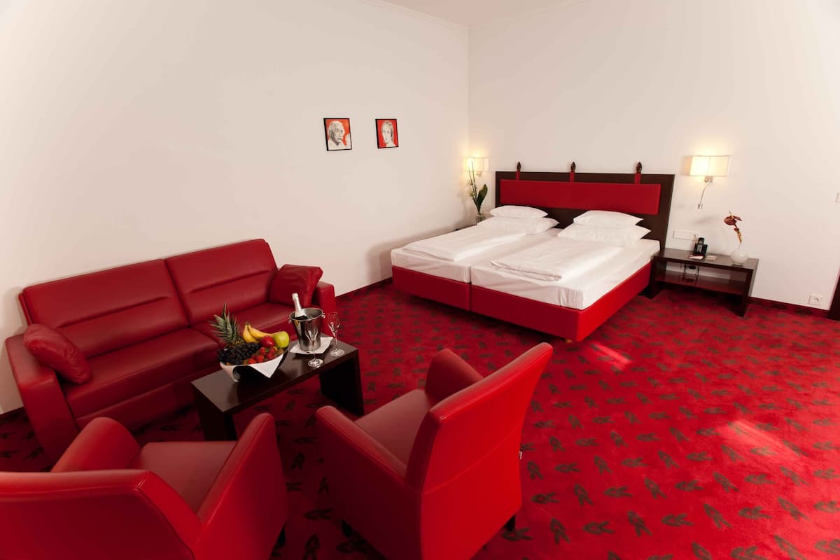Habitación de hotel con sofá rojo, cama con ropa de cama blanca, una mesita con fruta y una bebida, y una mesita de noche con una lámpara.