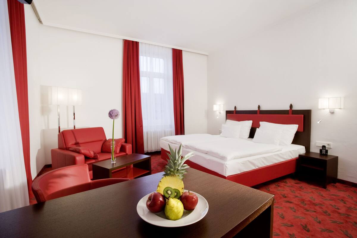 Habitación de hotel con cama, sofá rojo, mesa con fruta y alfombra roja.