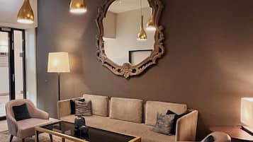 Ein modernes Wohnzimmer mit einem beigen Sofa, zwei Sesseln, einem Glastisch und einem großen, verzierten Spiegel.