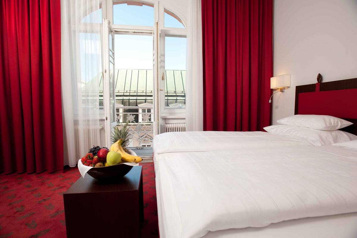 Habitación de hotel con cama, mesita de noche con frutero, cortinas rojas y vista a un edificio a través de una ventana abierta.