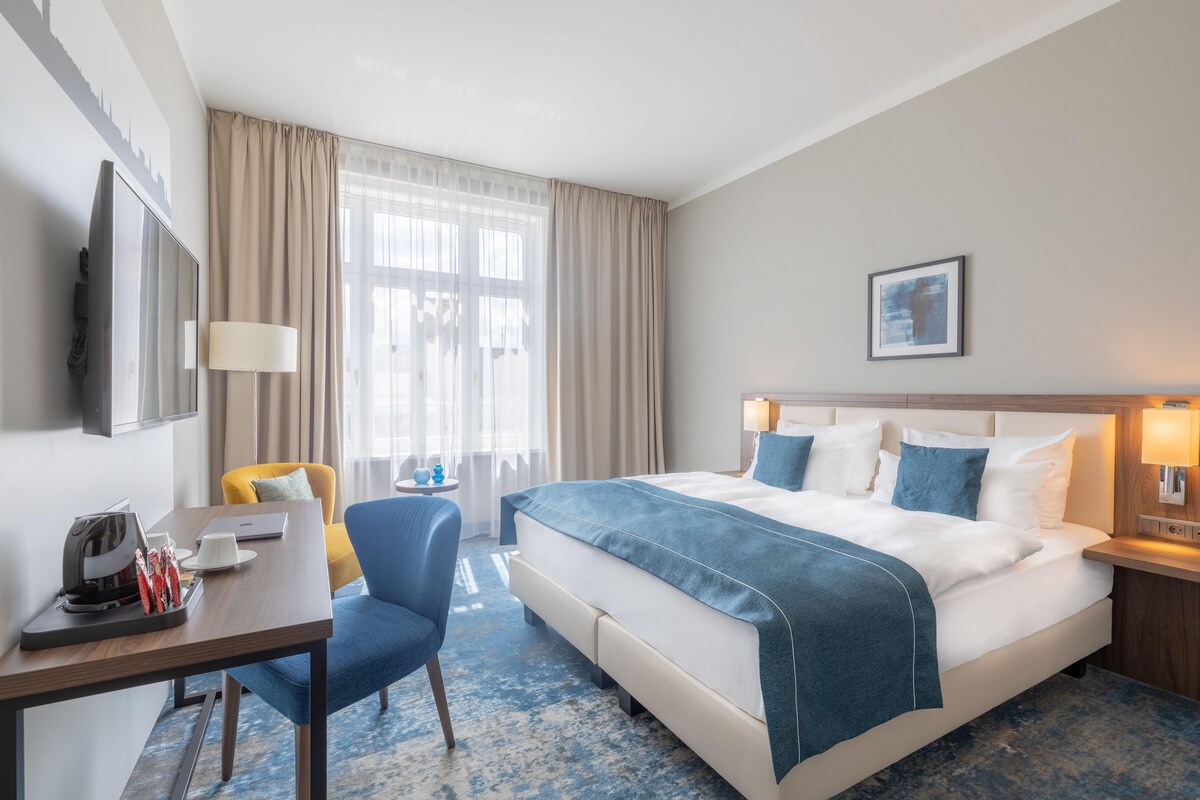 Quarto de hotel com uma cama grande, uma escrivaninha com cafeteira, uma cadeira e uma televisão.