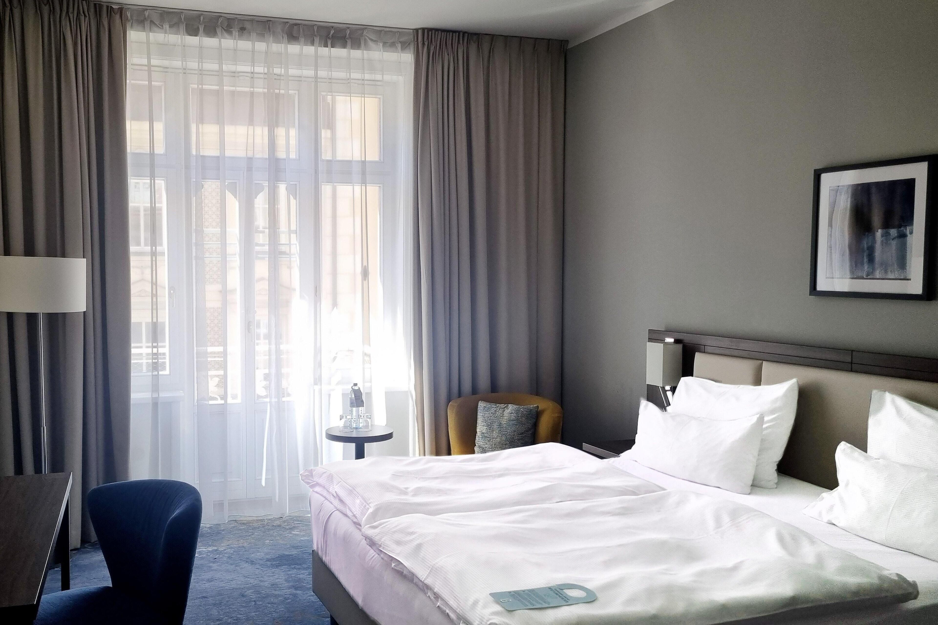 A hotel room with a bed, a chair, a small table, a lamp, and a window with curtains.