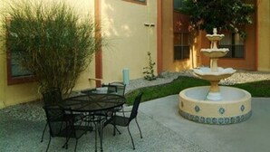 An outdoor patio with a round table and chairs, a decorative fountain, and a building with windows and a door in the background. - Pan American Inn & Suites (Albuquerque)