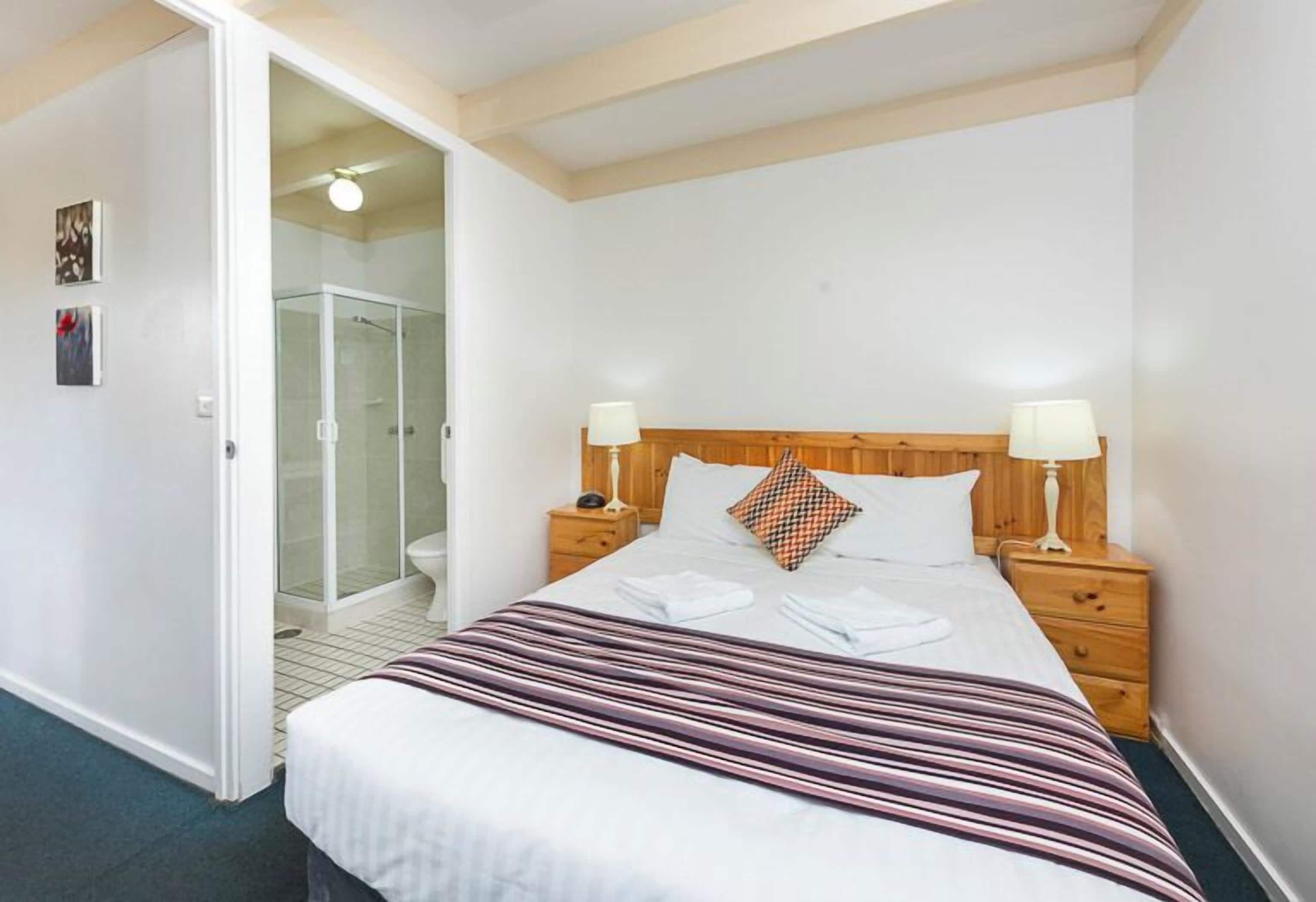 Habitació d'hotel amb llit, taules de nit, un espai per deixar coses al costat del llit i un bany amb dutxa de vidre, més una porta que dóna a una altra habitació.
