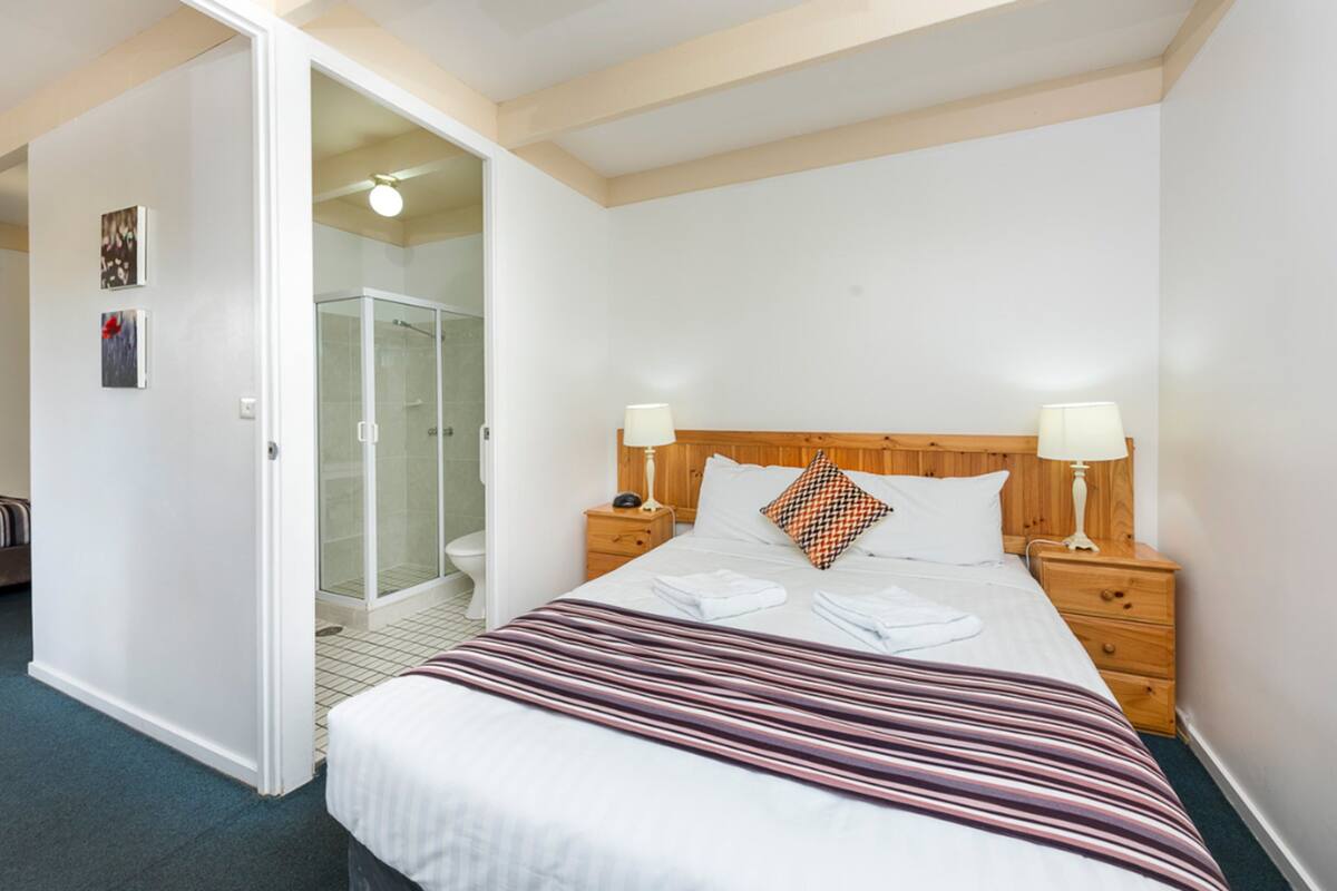 Une chambre d’hôtel comprenant un lit, deux tables de chevet avec des lampes, une tête de lit en bois, un espace douche et des toilettes.