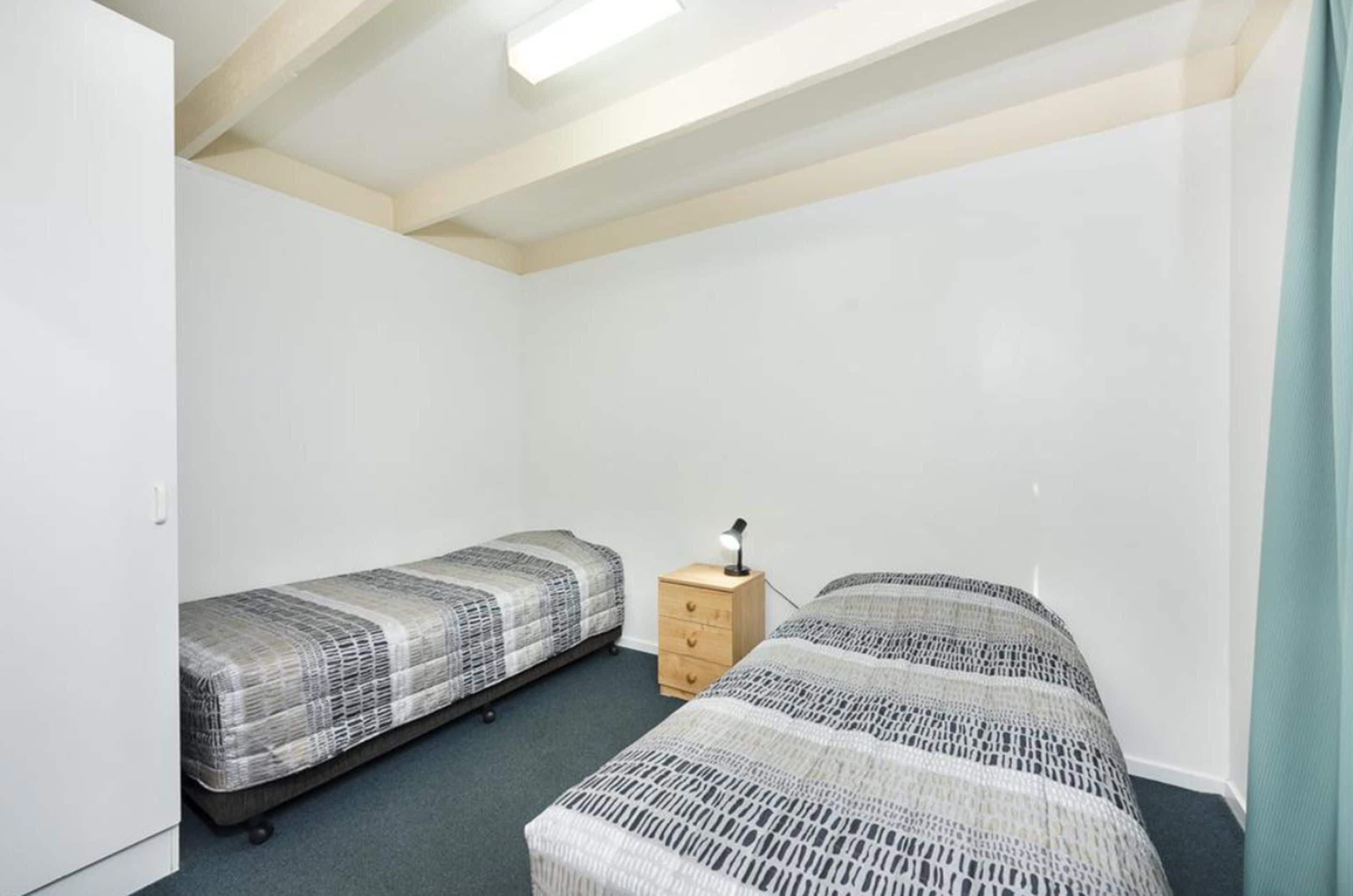 A small, clean room with two single beds, a wooden nightstand, and a desk lamp.