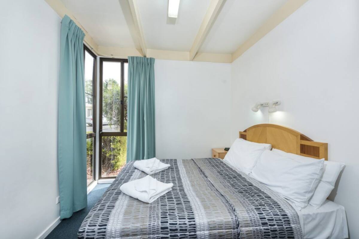 Une chambre à coucher avec un lit, une table de chevet, une fenêtre avec des rideaux et une vue sur la verdure à l’extérieur.