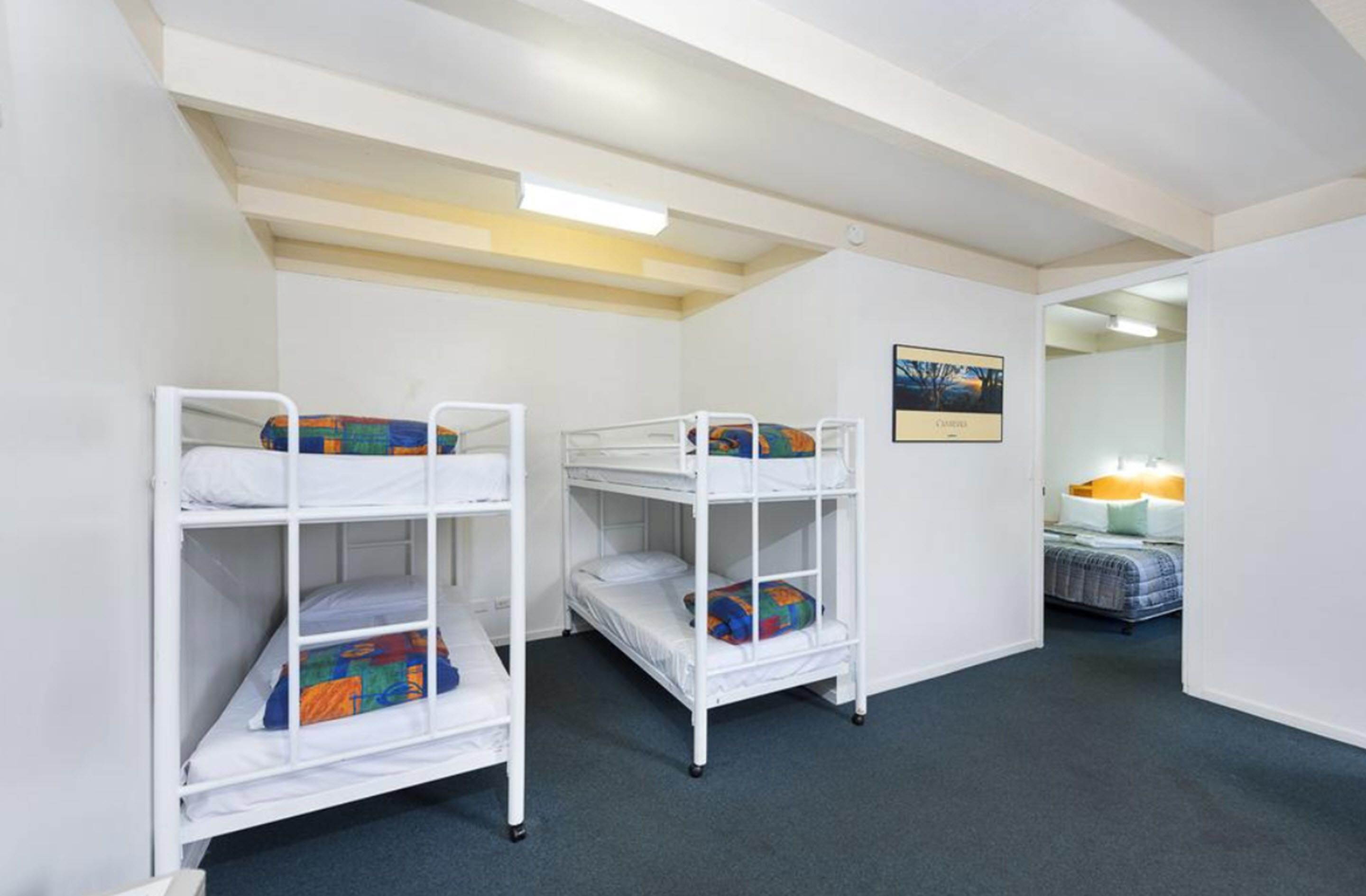 Ein Zimmer mit Etagenbetten, einem gerahmten Bild an der Wand und einem Bett mit Lampe im Hintergrund.