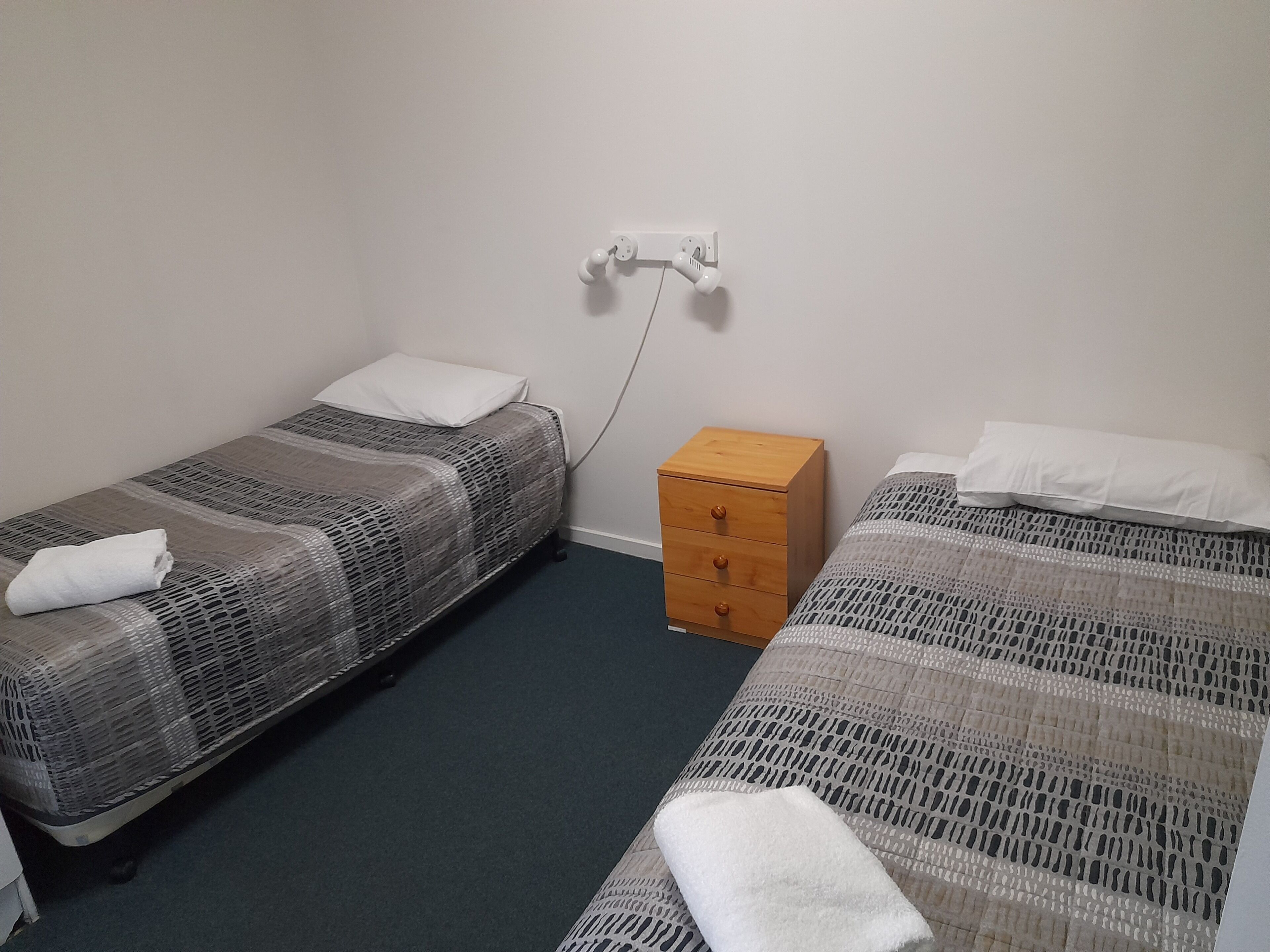 Une chambre avec deux lits simples, une table de chevet en bois et une prise électrique au mur.