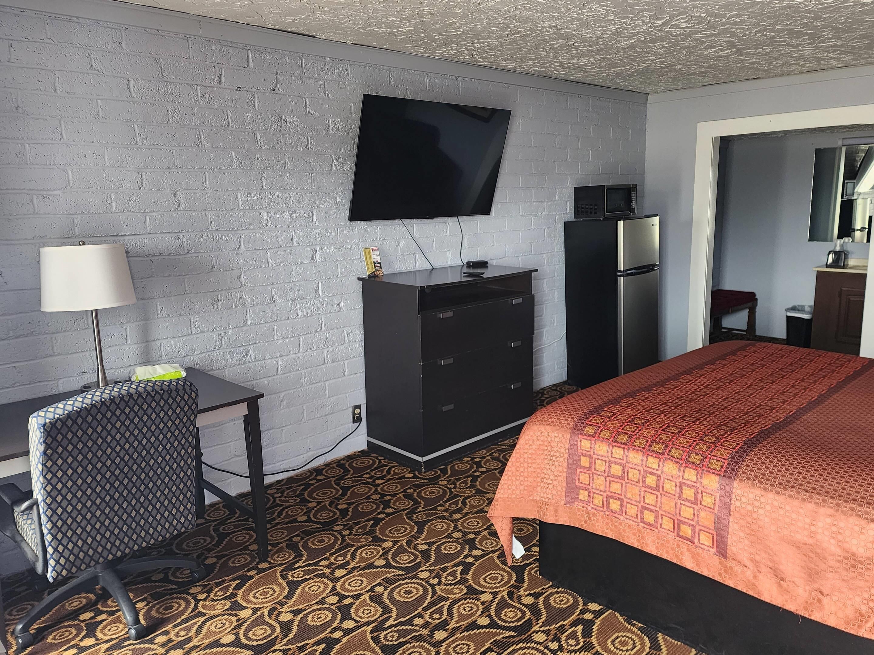 Une chambre d’hôtel comprenant un lit, un bureau avec une chaise, un téléviseur fixé au mur, un réfrigérateur et une moquette à motifs.