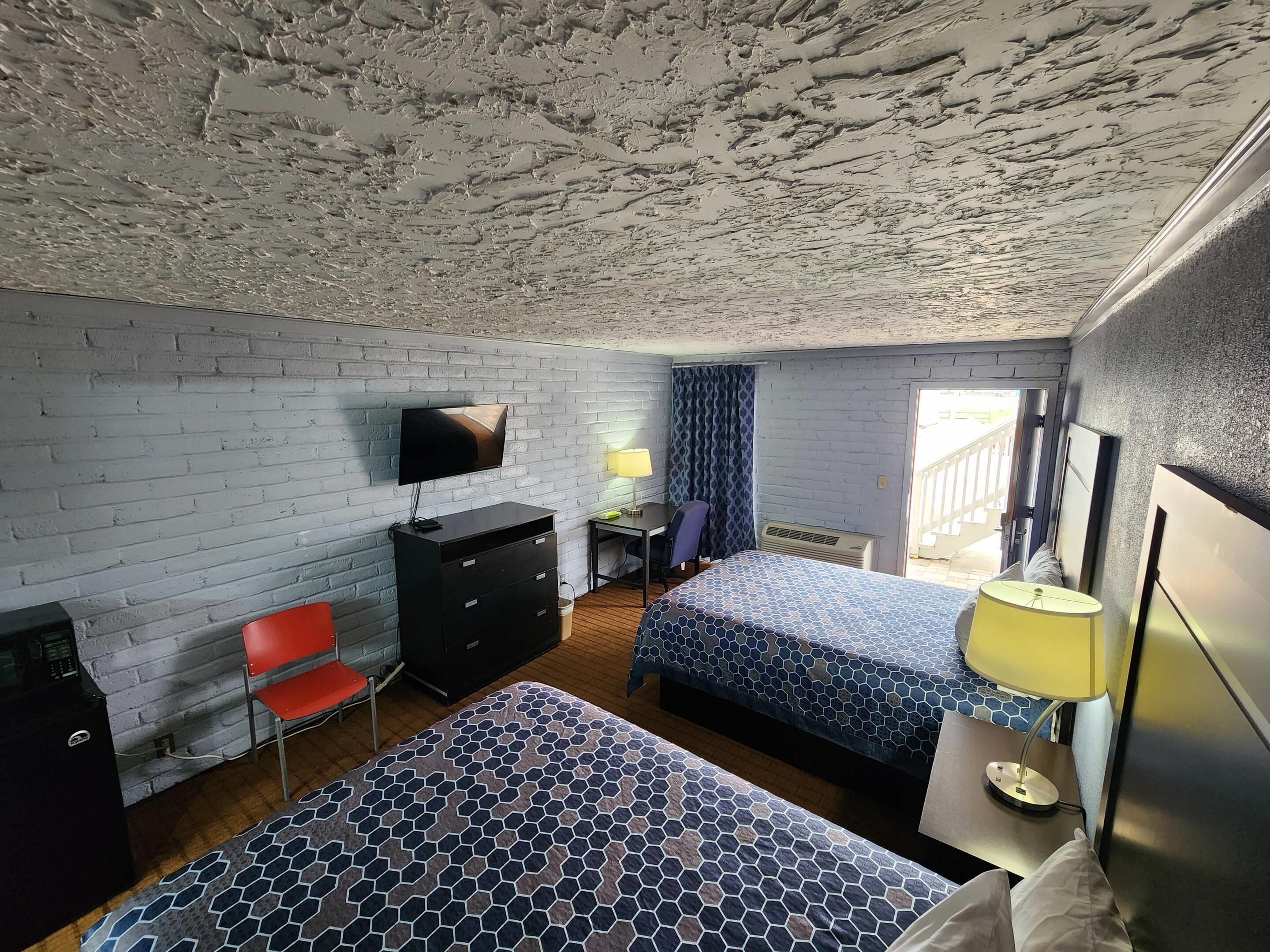 Una habitación con pared de ladrillo, un televisor montado en la pared, un escritorio con una silla, una cama con colcha estampada y una lámpara en la mesita de noche.