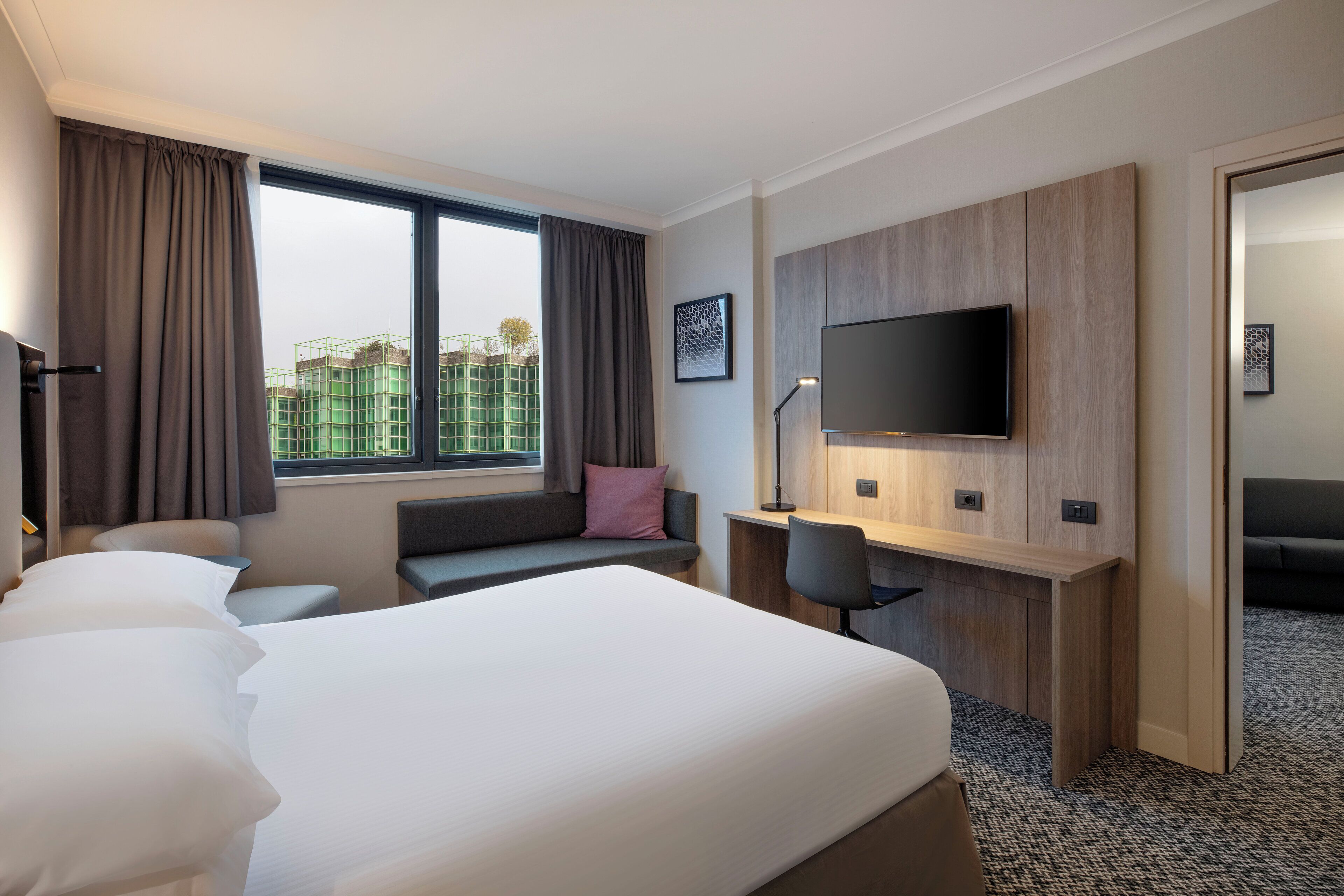 Une chambre d’hôtel équipée d’un lit, d’un bureau, d’une chaise, d’une télévision et d’une grande fenêtre.