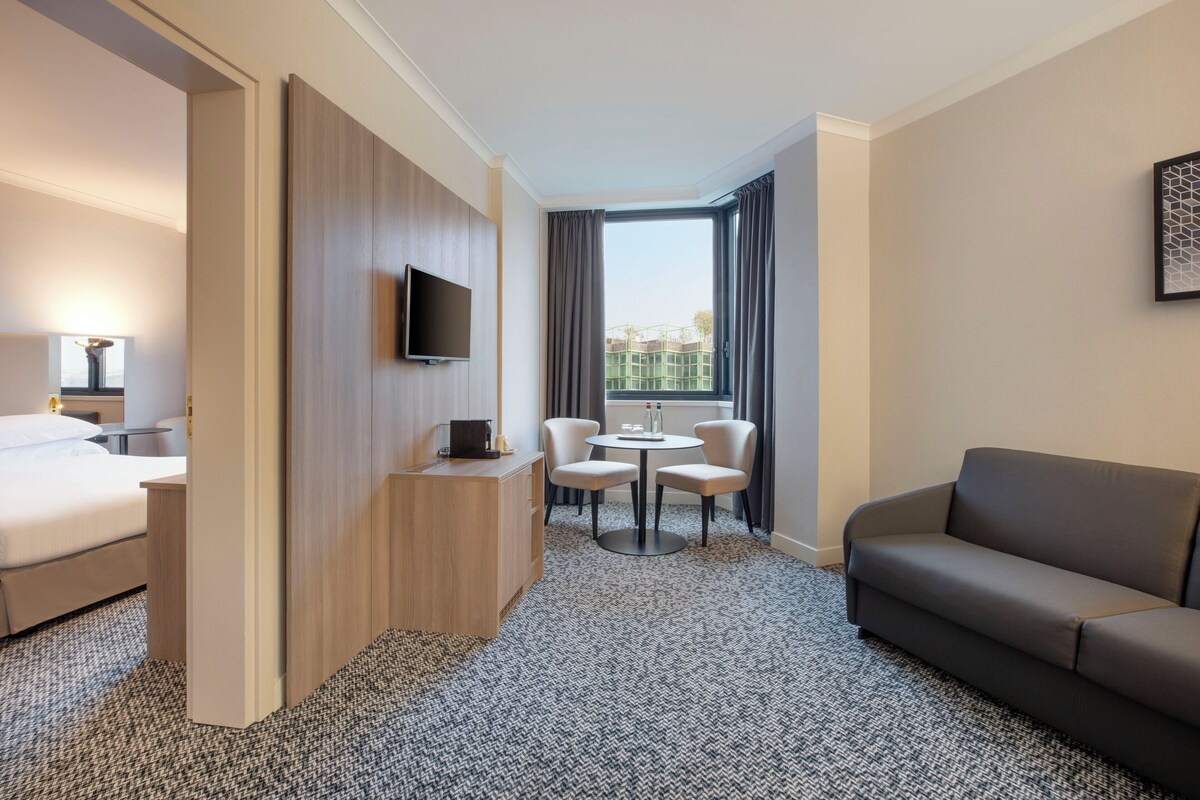 A hotel room with a bed, a TV, a desk, a sofa, a small table with chairs, and a window with curtains.