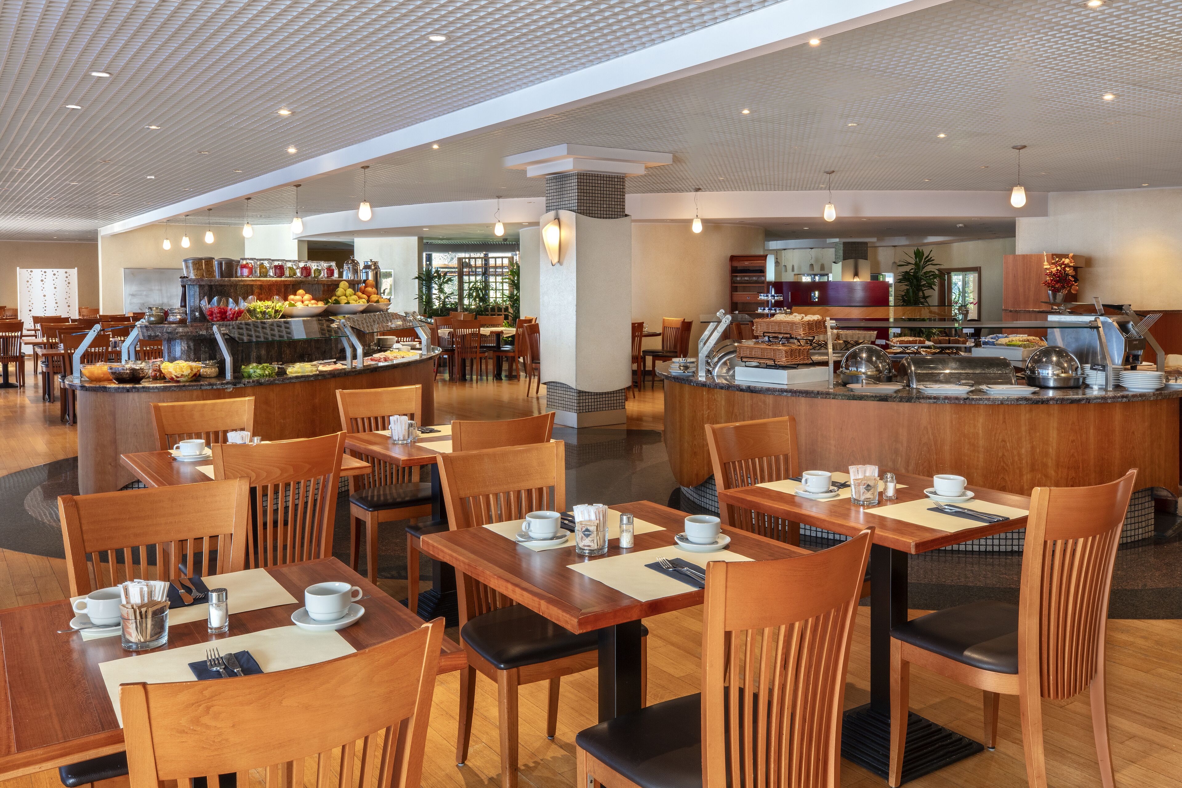 Un comedor moderno con mesas y sillas de madera, una barra de buffet con diversos alimentos y un interior limpio y bien iluminado.