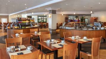 Un comedor moderno con mesas y sillas de madera, una barra de buffet con diversos alimentos y un interior limpio y bien iluminado.